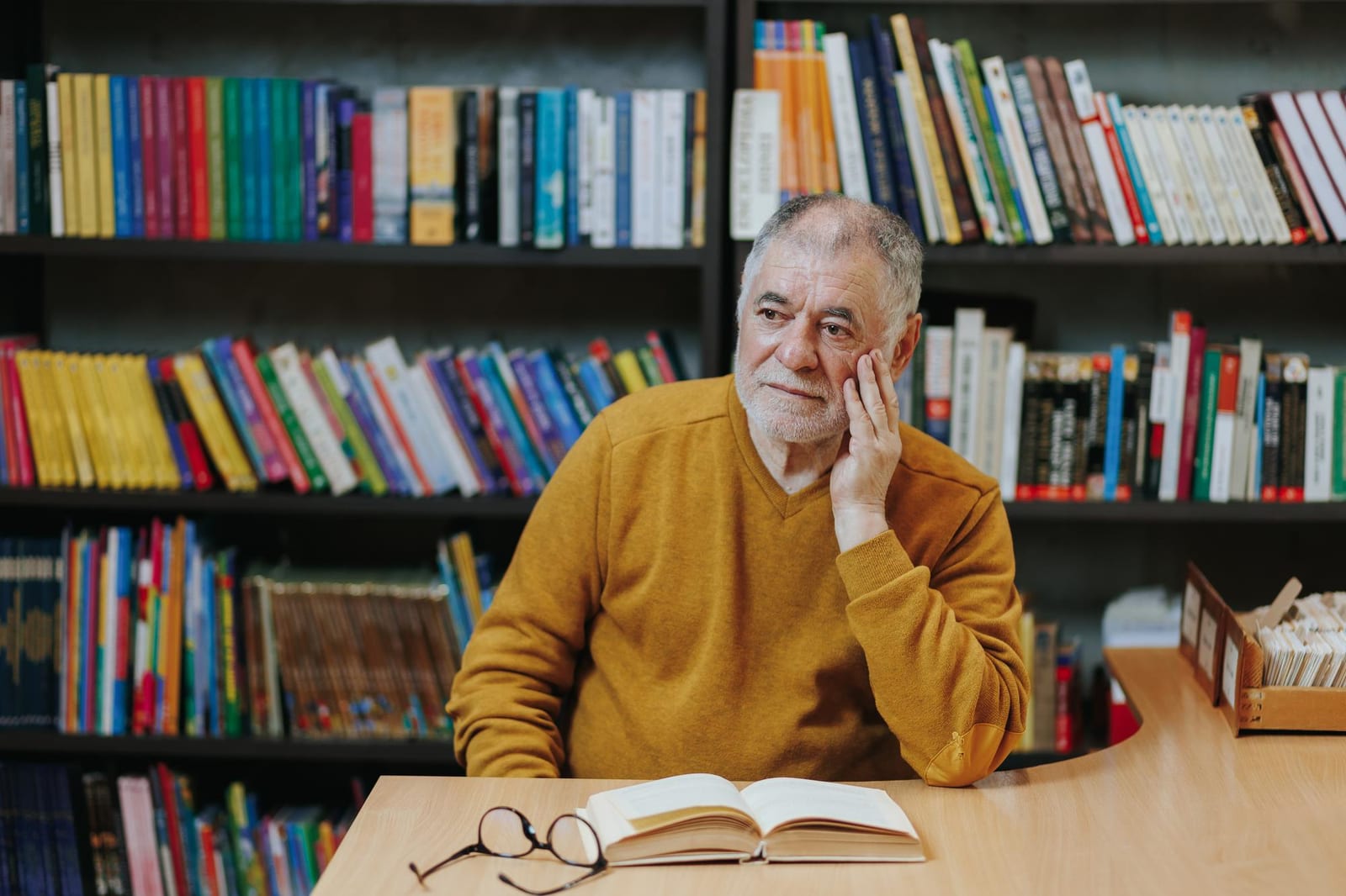 Homem sentado com um livro aberto, refletindo, diante de estantes coloridas em uma biblioteca