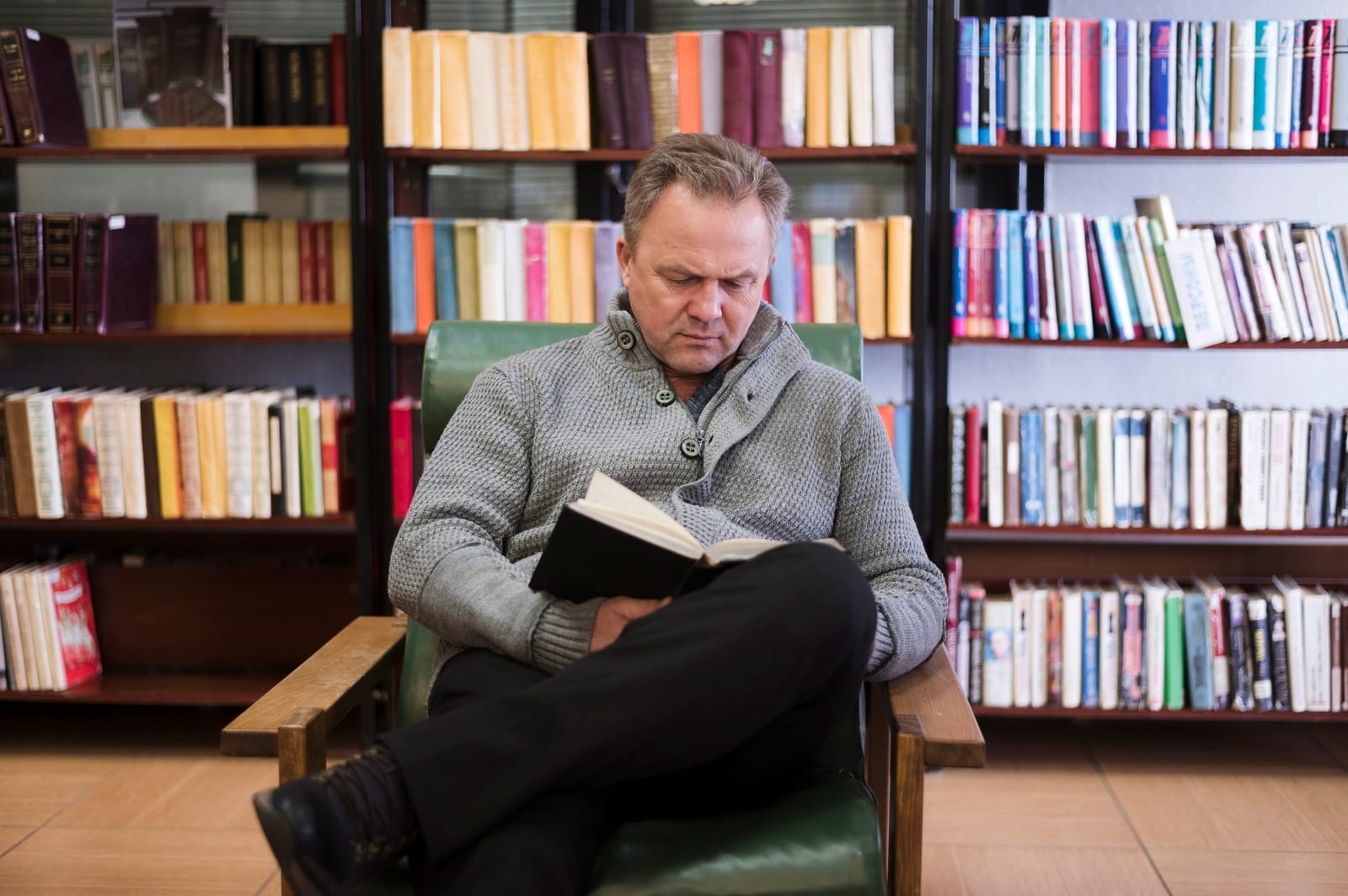 Homem lendo um livro em poltrona de couro verde diante de estantes repletas de livros coloridos