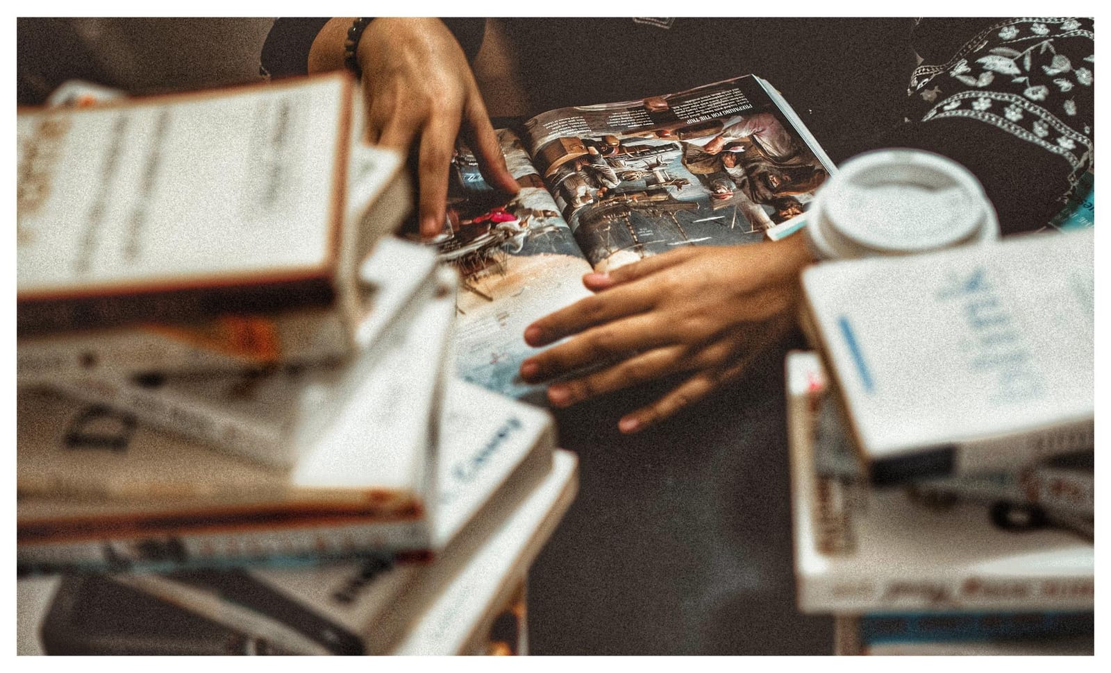 Mãos folheando um livro aberto sobre uma pilha de livros, com uma xícara de café ao lado